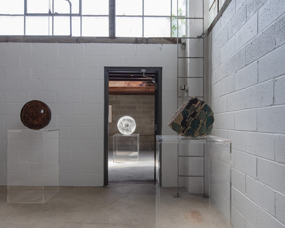 Art gallery featuring three abstract sculptures on clear pedestals in an industrial-style setting with concrete walls.