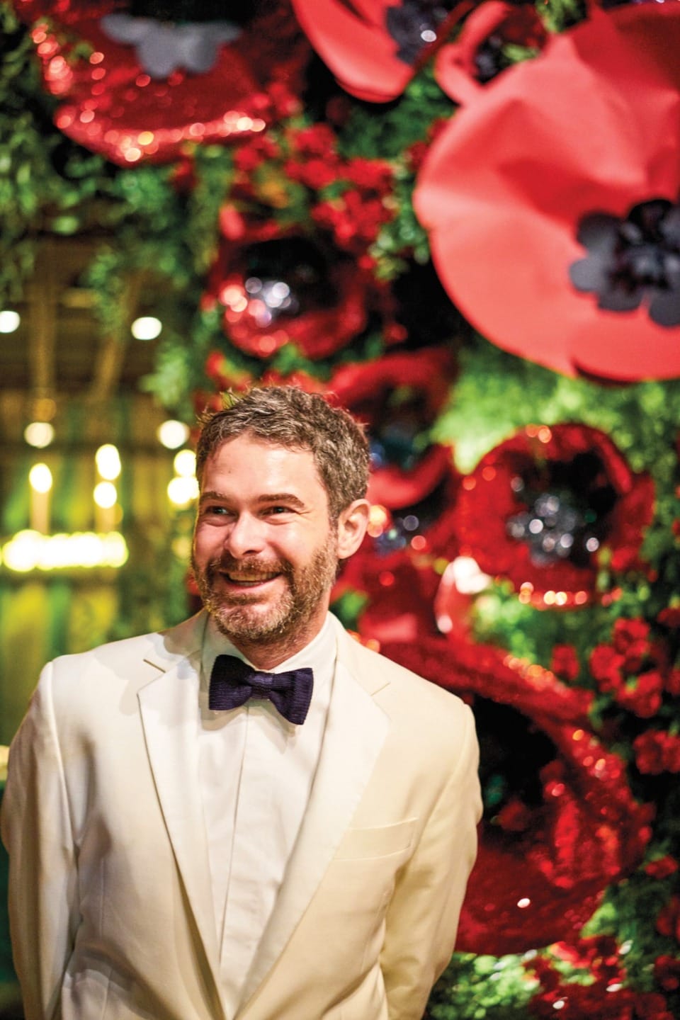 Man in a white suit with a bow tie smiling in front of a large red poppy flower display.