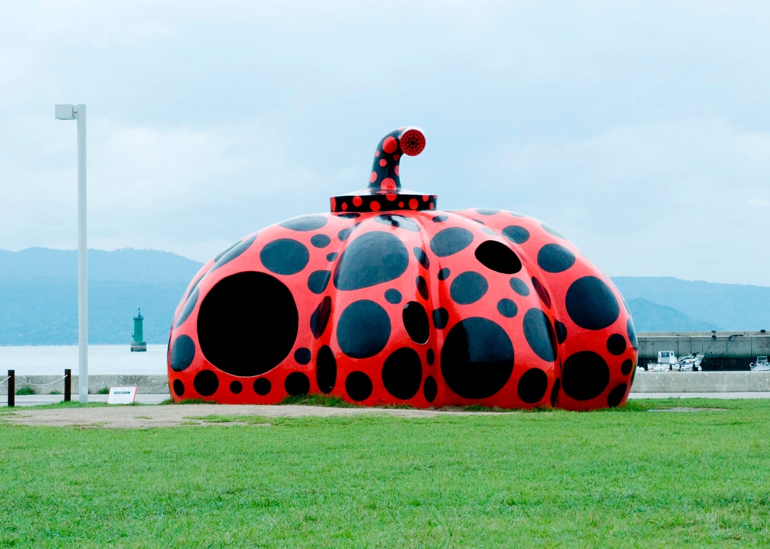 Red and black polka-dotted pumpkin sculpture on a grassy area near a waterfront with hills in the background.