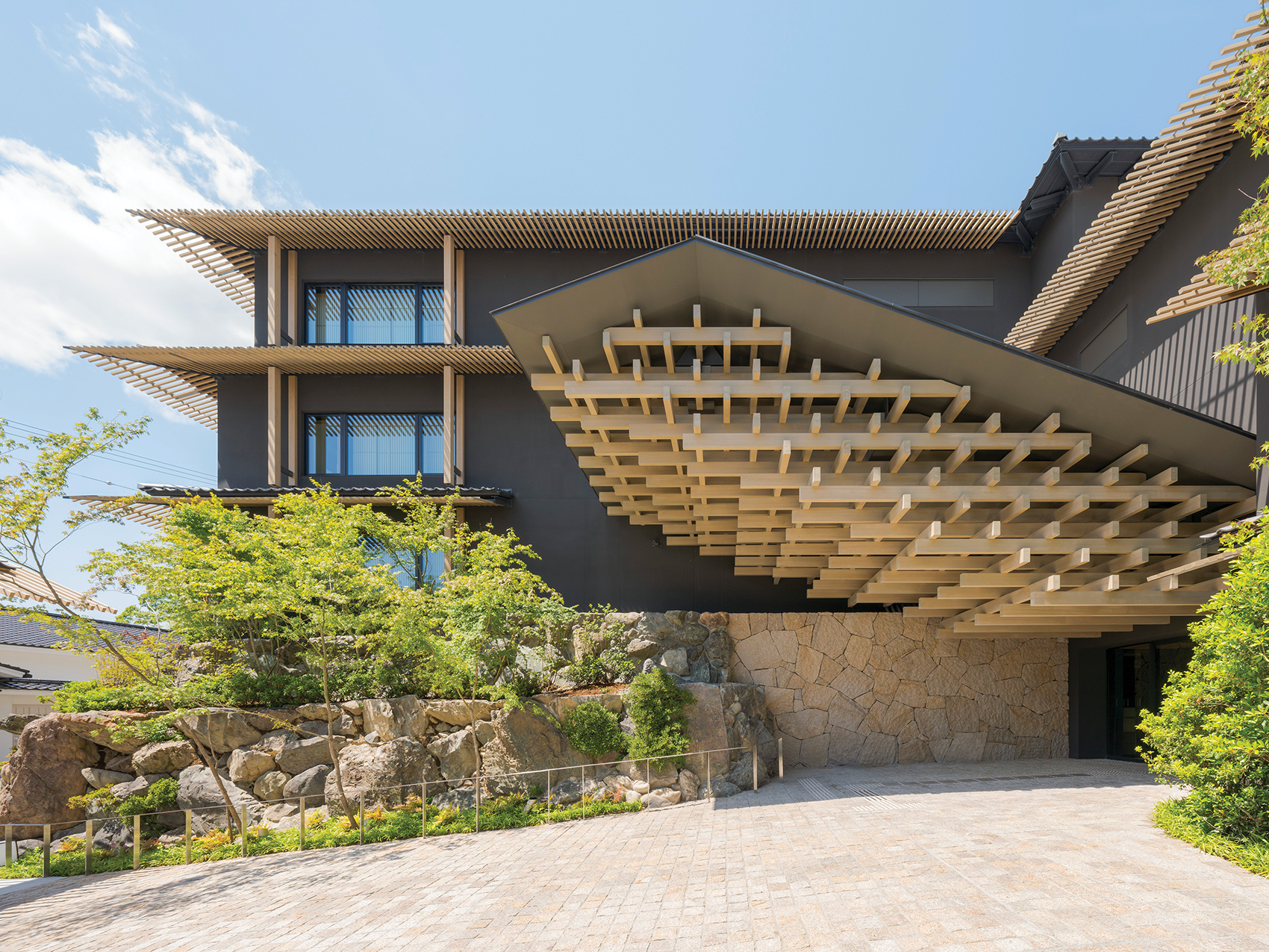 Modern building with unique wooden architectural design, surrounded by greenery and stone elements, under a clear blue sky.