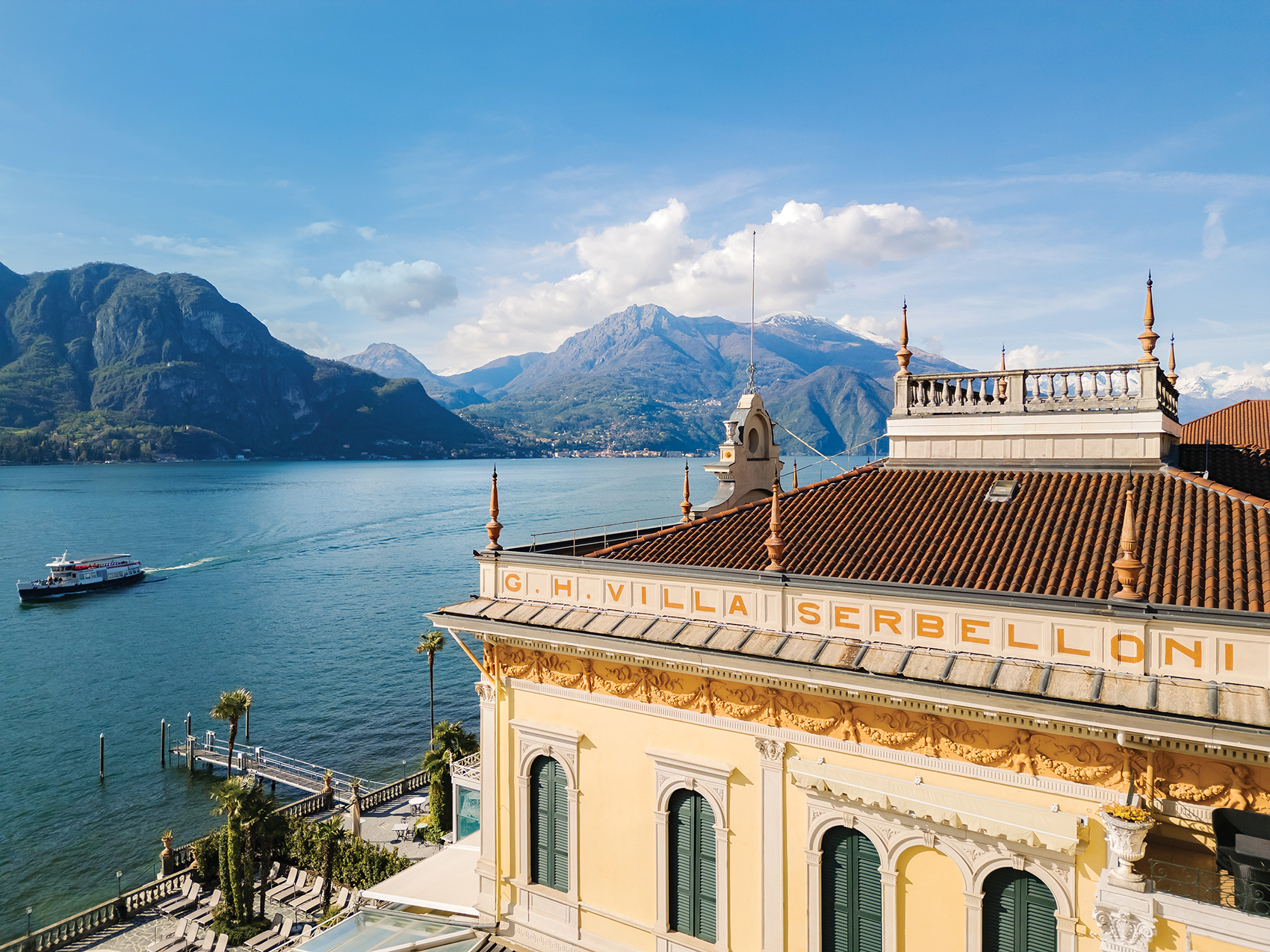 Historic villa overlooking Lake Como with a boat passing by, framed by lush mountains and a clear blue sky in the background.