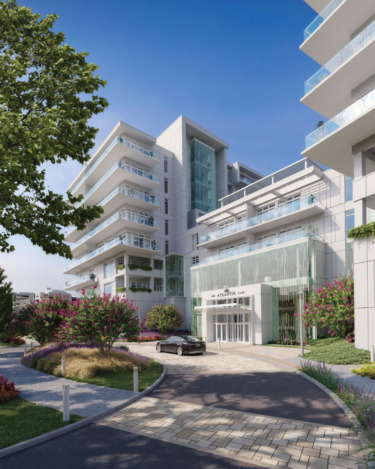 Modern apartment building with glass balconies, surrounded by landscaped gardens and a driveway entrance under a clear blue sky.