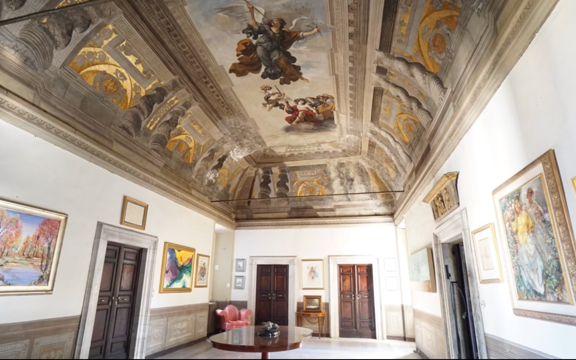 Ornate room with decorated ceiling, paintings on walls, wooden doors, and a round table in center.