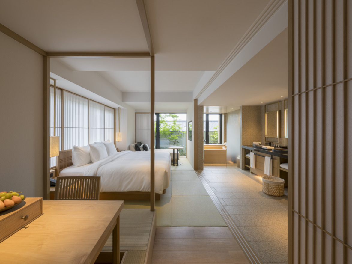 Modern hotel room with double bed, wooden furniture, large windows, and an open bathroom with a garden view.
