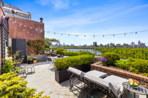 Rooftop terrace with seating, string lights, and lush greenery, offering a scenic view of a city skyline over a park.