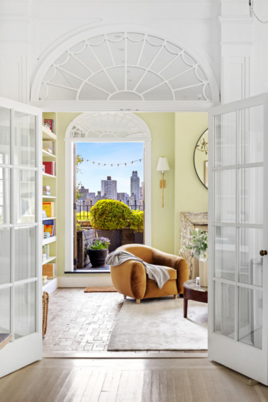 Bright living room with an arched window view of cityscape, featuring a cozy armchair, plants, and shelves with books.