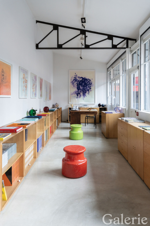 Modern art gallery with colorful stools, framed artwork on walls, and books on shelves under large windows.