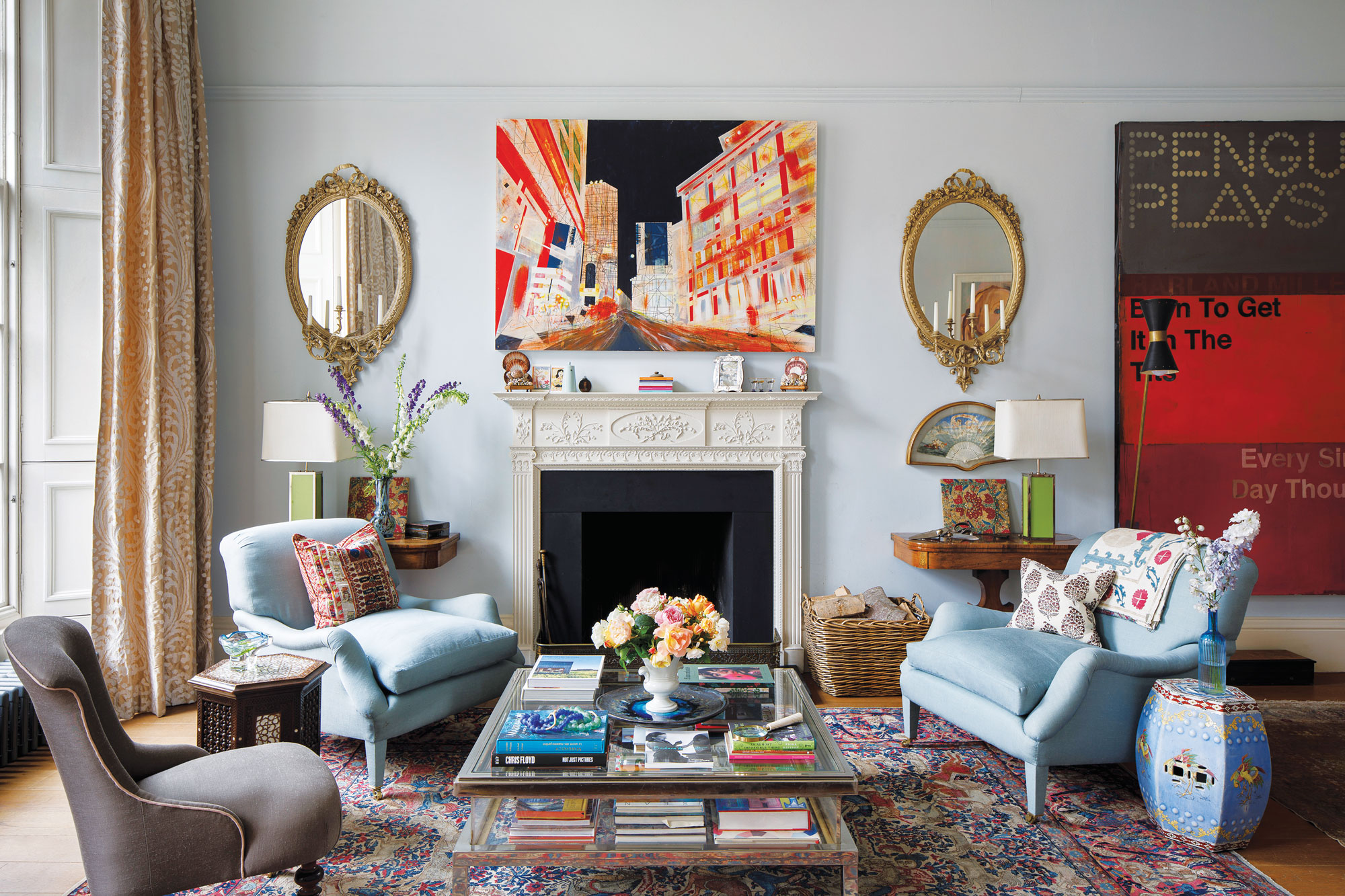 Eclectic living room with colorful art, ornate mirrors, blue armchairs, and a patterned rug in front of the fireplace.