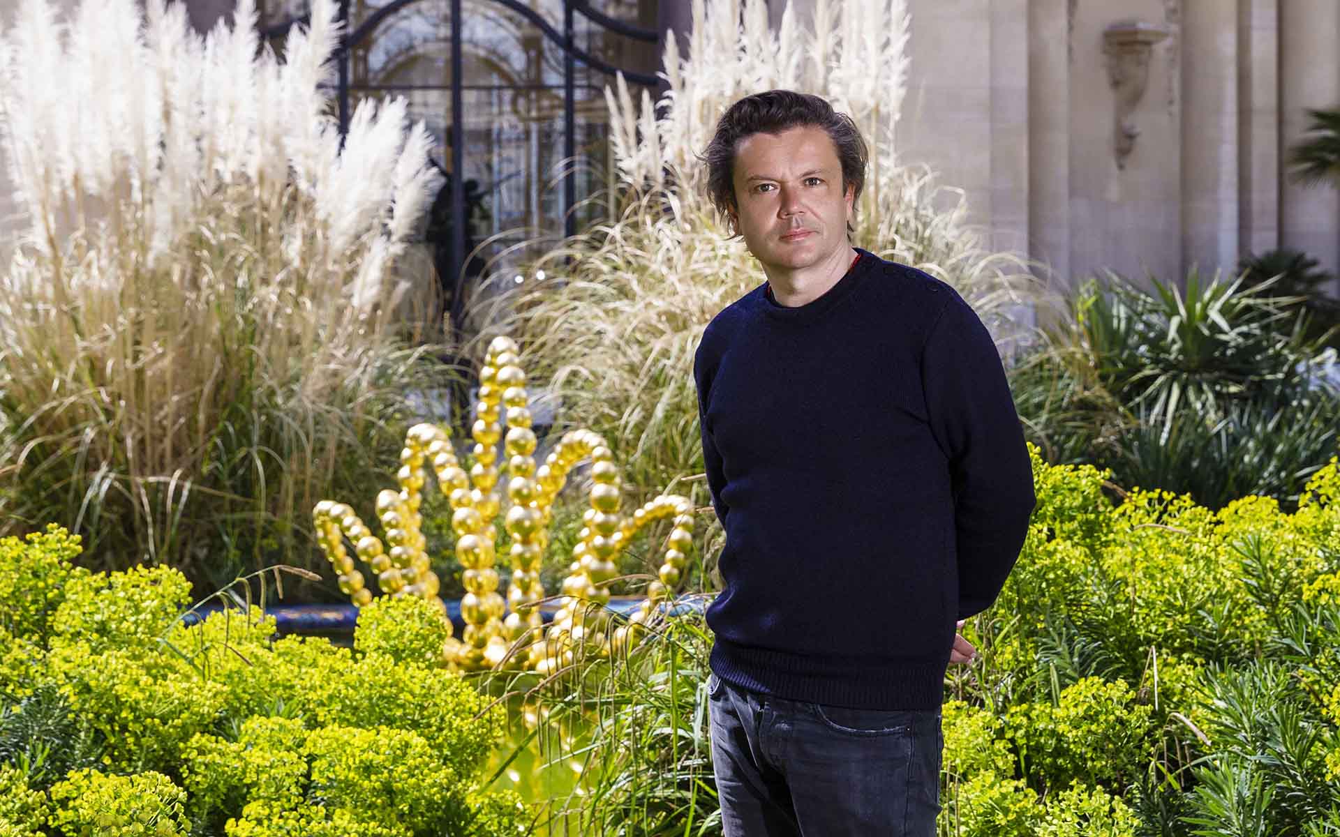 Person standing in a garden with ornamental plants and a decorative golden structure in the background.