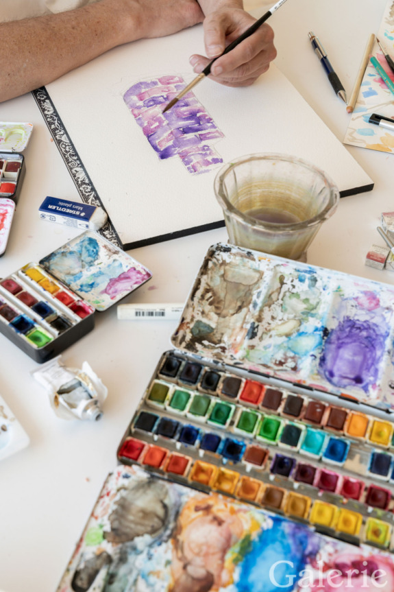 Artist painting with watercolors using purple hues on canvas surrounded by colorful paints and a glass of water.