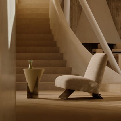 Modern minimalist living room with beige lounge chair, geometric side table, and curved staircase in warm lighting.