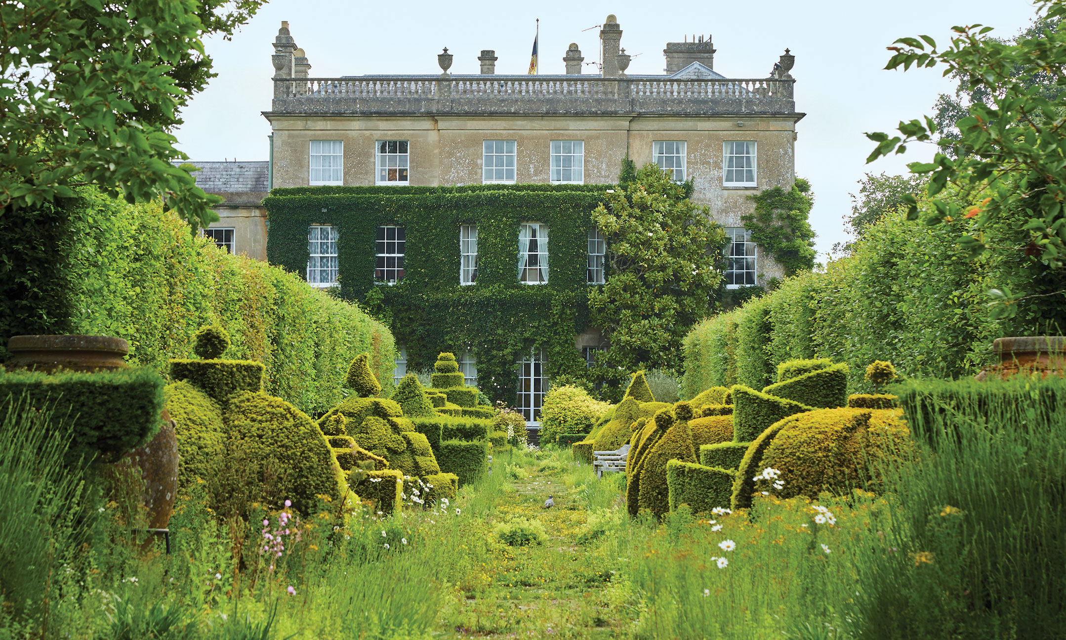Manor house with lush green gardens, topiary forms, and a variety of flowers, creating a serene and picturesque scene.