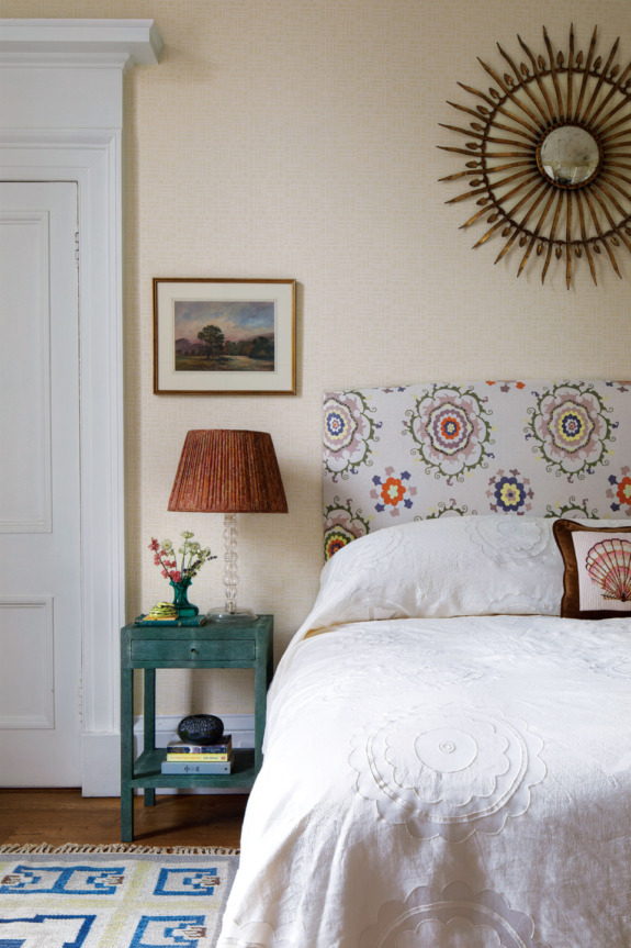 Cozy bedroom interior with floral headboard, sunburst mirror, colorful nightstand, and textured white bedspread.
