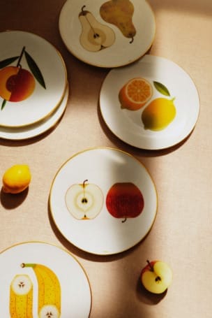 Ceramic plates with fruit illustrations, including pear, orange, apple, and banana, arranged on a beige surface.