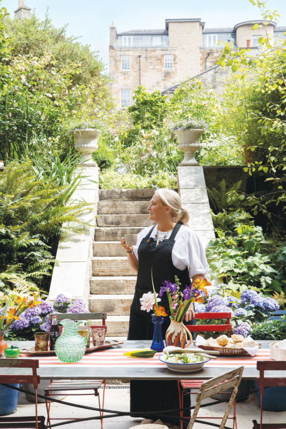 Woman standing in a lush garden by a set outdoor table with flowers, plants, and a stone staircase in the background.