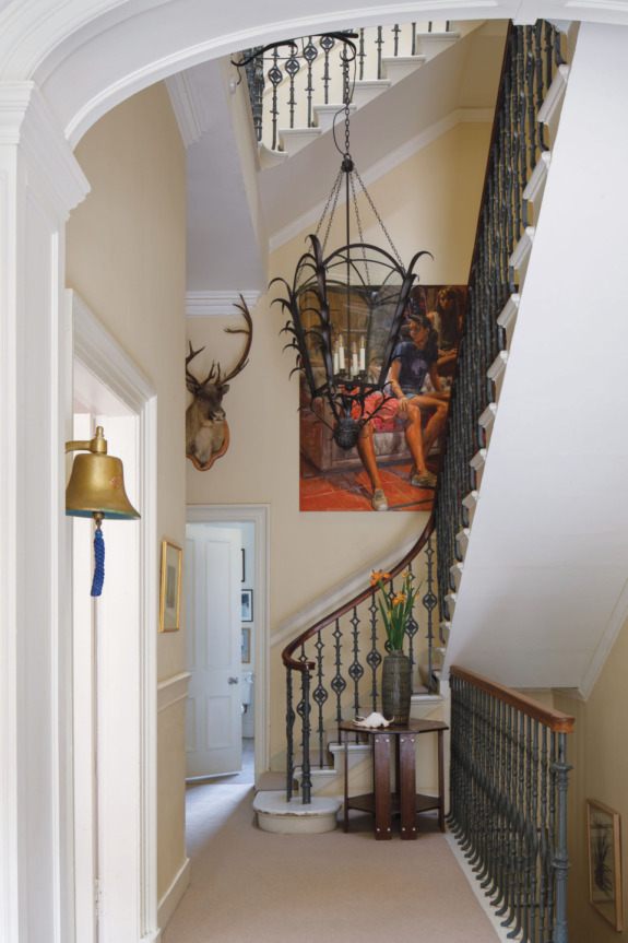 Spiral staircase in a narrow hallway with a large painting, mounted deer head, and decorative bell on the wall.