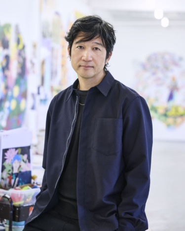 Person in a dark shirt standing in an art studio with colorful artwork in the background.