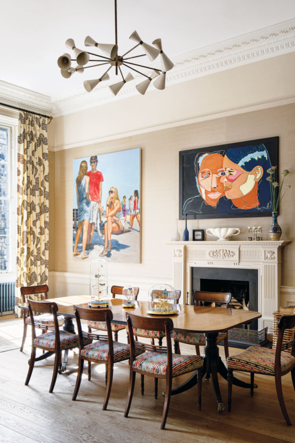 Dining room with a wooden table, colorful chairs, modern chandelier, and artwork on cream walls above a decorative fireplace.