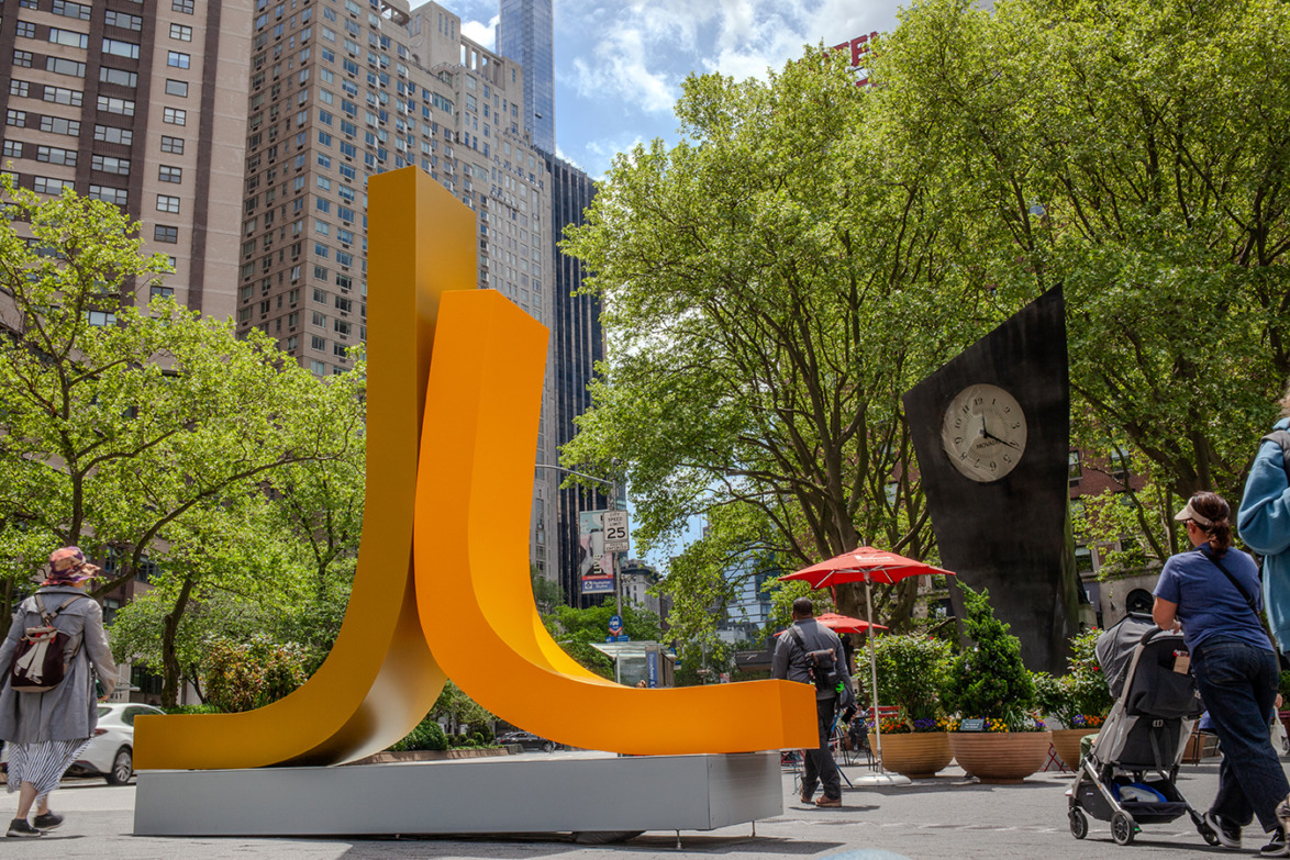 Bright orange sculpture in a bustling urban park setting with people walking and lush green trees around.