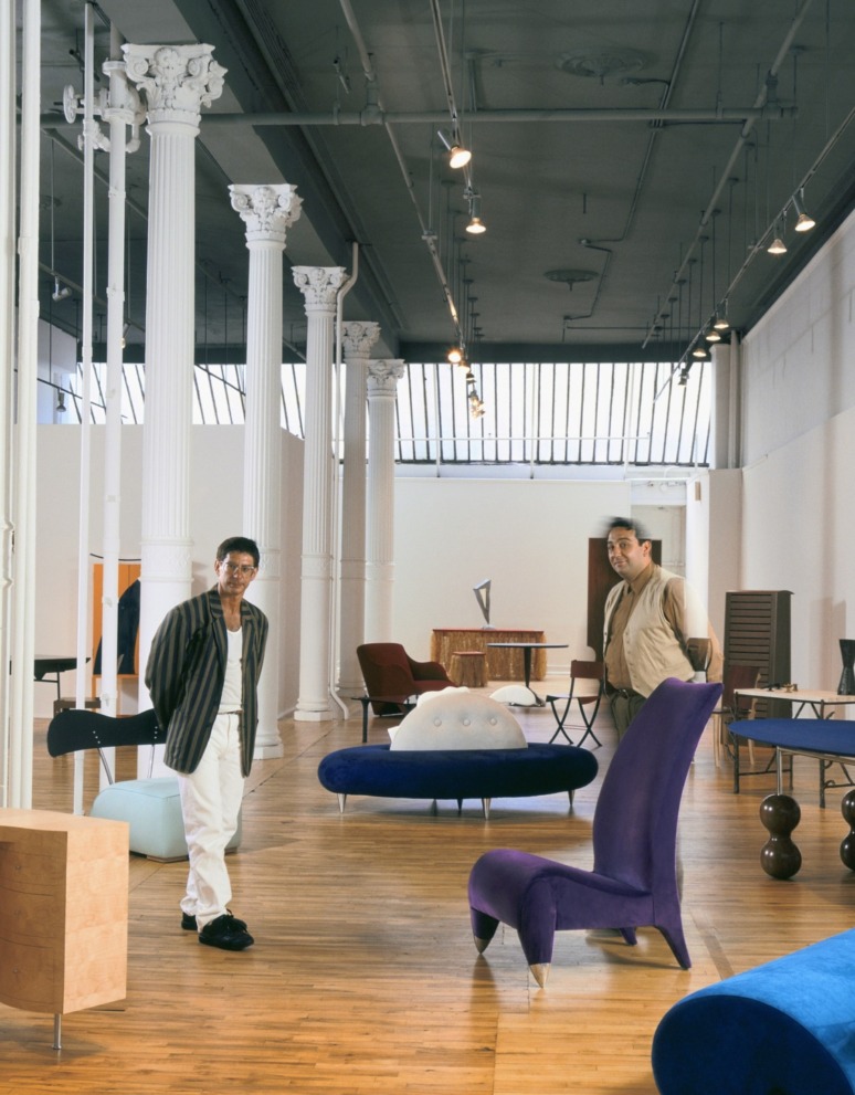 Two people standing in a modern furniture showroom with assorted colorful chairs and decorative columns.