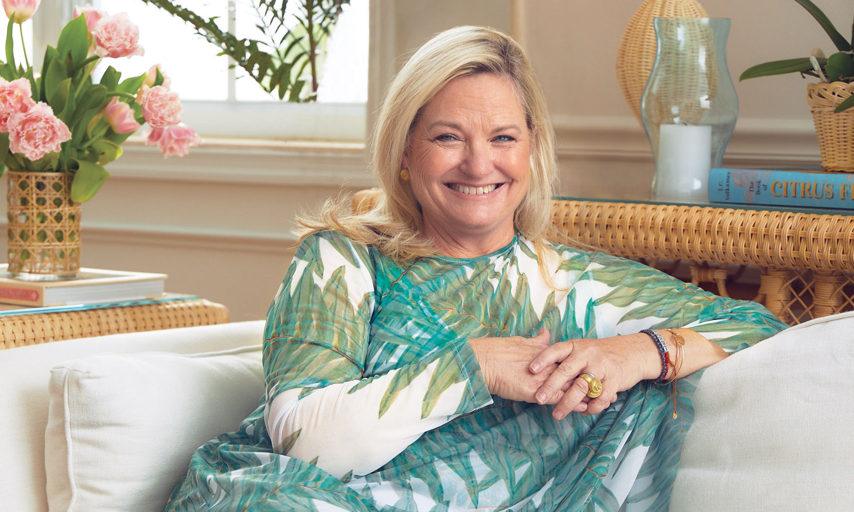 Smiling person seated on a sofa with a plant-patterned outfit, surrounded by flowers and books in a bright room.