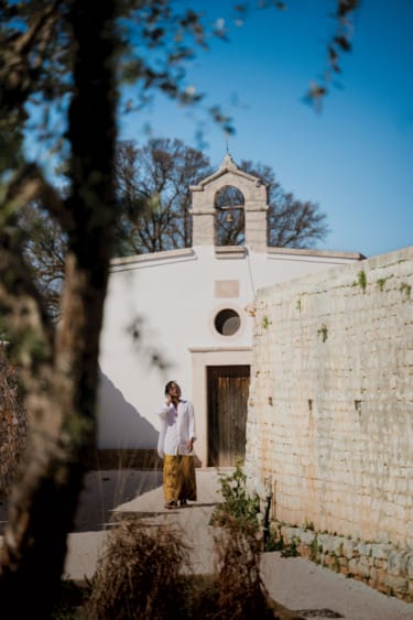Person in yellow pants and white shirt standing near a stone wall and small church under a clear blue sky.