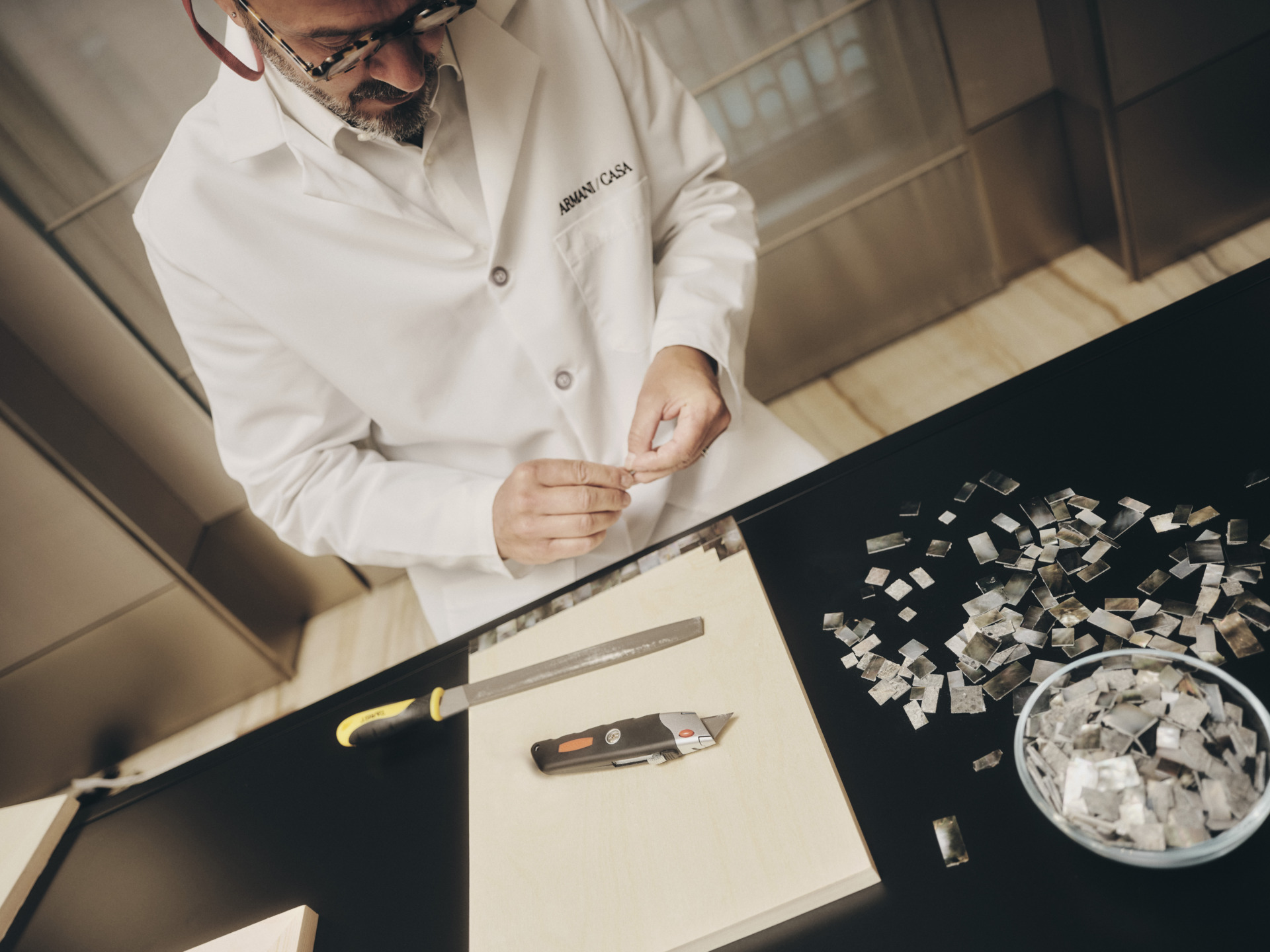 A person in a white lab coat carefully arranging small tiles on a black table with a utility knife nearby.