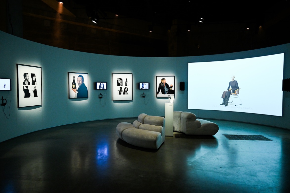 Modern art gallery with portraits displayed on curved wall, large digital screen, and unique gray seating in center.