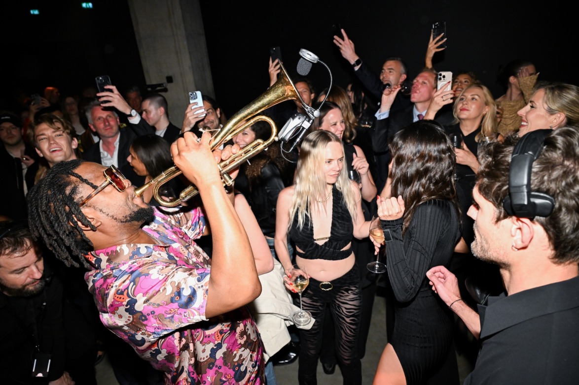 Musician playing trumpet in a lively party with people dancing and taking photos in the background.