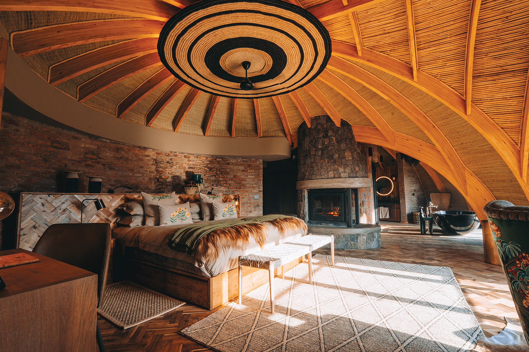 Cozy round bedroom with wood ceiling, stone fireplace, and bohemian decor, featuring a large bed and wicker accents.