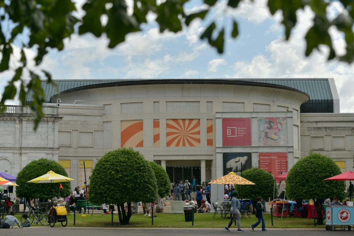 People visit an art museum with colorful banners and outdoor activities, surrounded by trees and puffy clouds in the sky.
