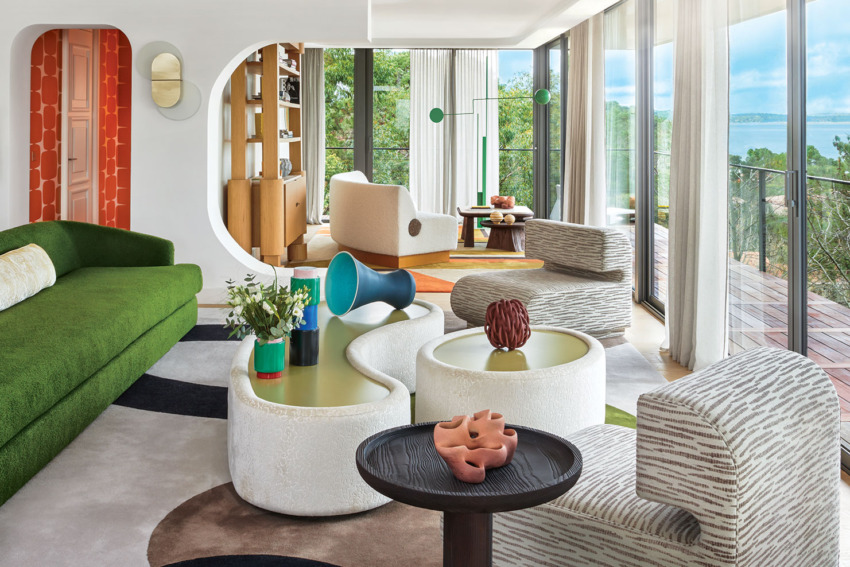 Modern living room with green sofa, patterned chairs, colorful decor, large windows, and a view of trees and water.