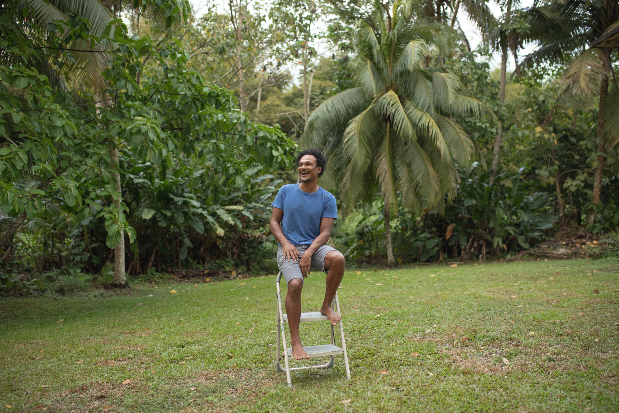 Man sitting on a step ladder in a tropical garden, surrounded by lush green trees and grass, smiling and looking upward.