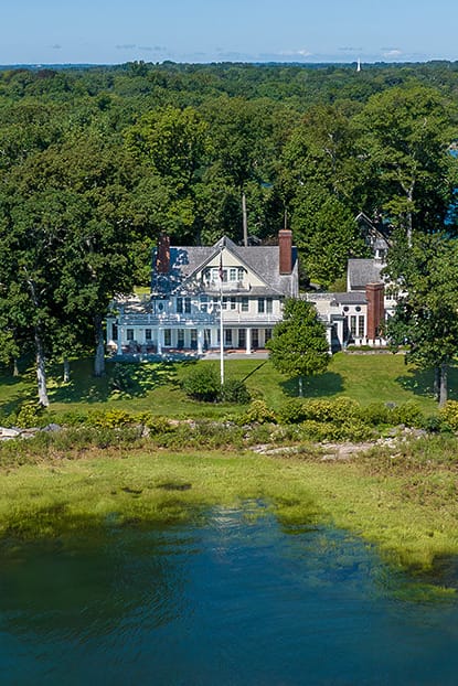 Aerial view of a secluded lakeside mansion surrounded by lush trees and rocky shoreline under a clear blue sky.
