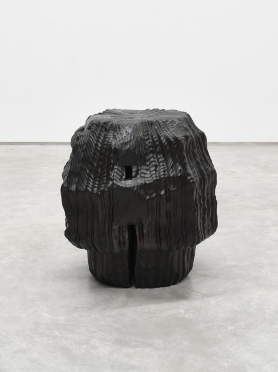 Sculptural black wooden stool with a textured surface resembling a tree trunk set on a gray concrete floor.