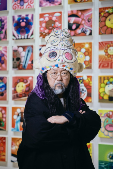 Artist in a colorful patterned hat stands in front of a vibrant wall of art with playful, colorful designs