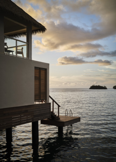 Overwater bungalow with steps and a ladder leading into the ocean at sunset, surrounded by calm water and distant island.