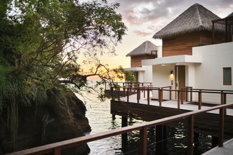 Overwater bungalows with thatched roofs at sunset near rocky shoreline and lush vegetation