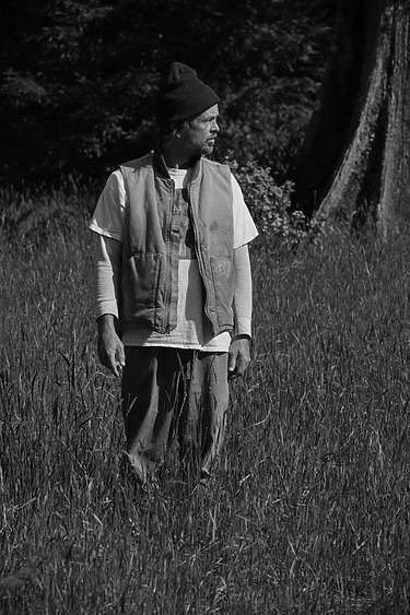 A person wearing a beanie and vest stands in a grassy field looking to the side, with trees in the background.