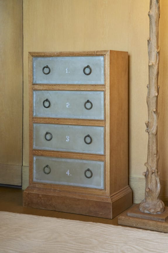 Wooden four-drawer cabinet with numbered metal front panels and round handles, beside a tree-like carved lamp.