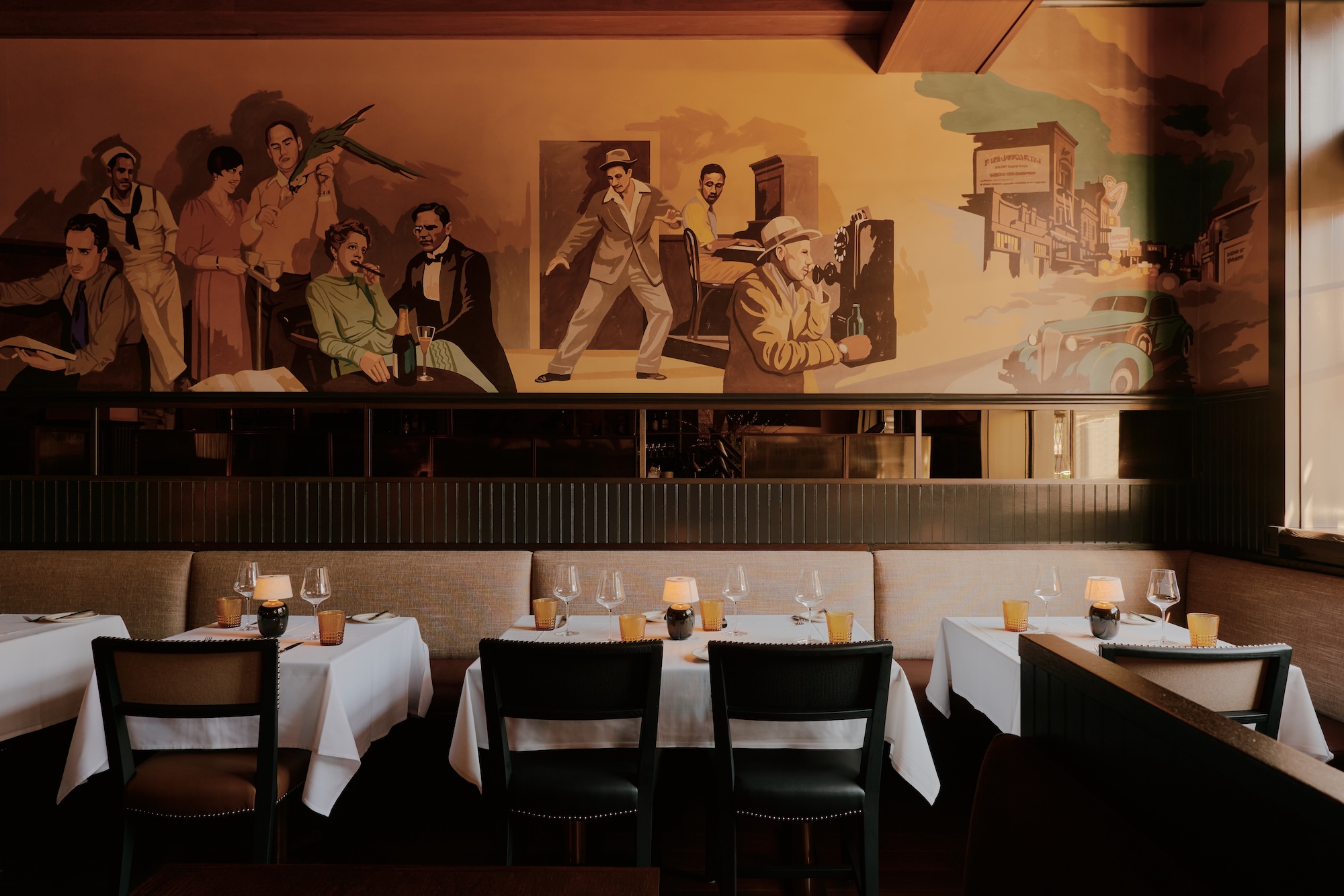 Cozy restaurant with a vintage mural and neatly set tables, featuring glasses, candles, and a warm ambiance.