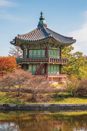Traditional Korean pavilion by a pond with vibrant autumn foliage and a distant pagoda under a clear blue sky.