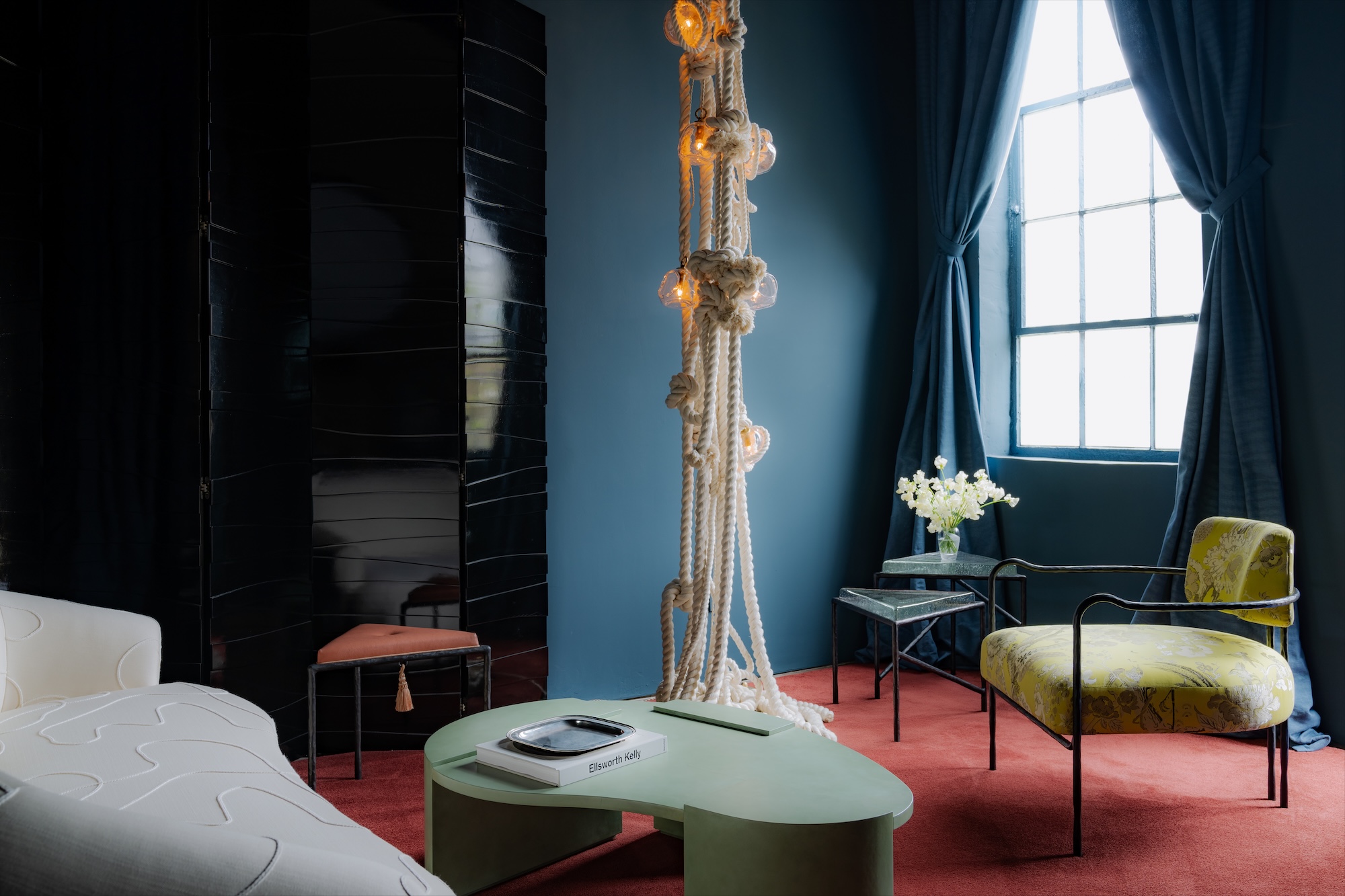 Modern living room with eclectic furniture, blue walls, floor-to-ceiling window, red carpet, rope lamp, and decorative accents.