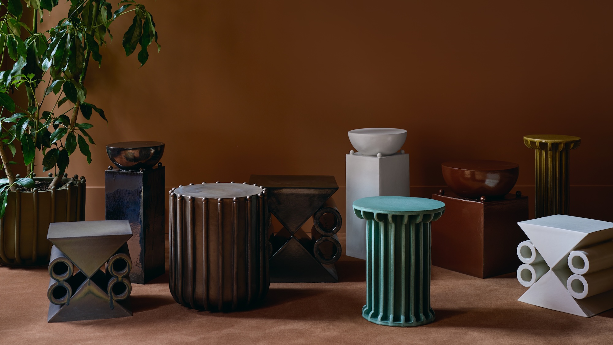Assorted modern ceramic stools on a brown carpet with a green plant in the corner against a warm brown wall.