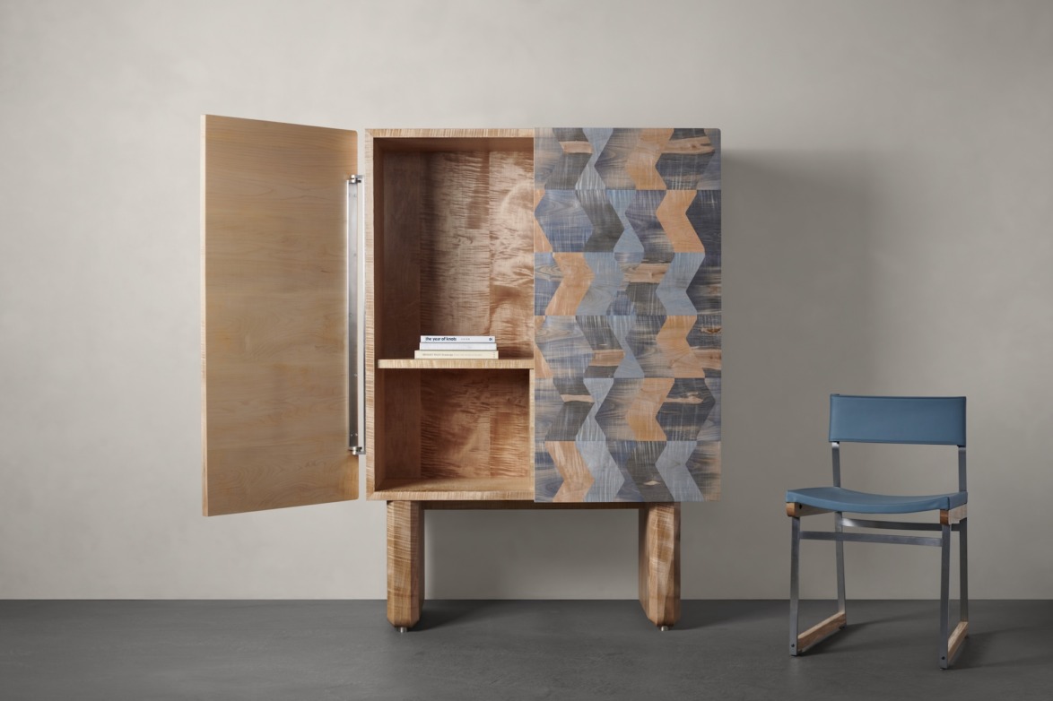 Wooden cabinet with geometric patterned door sits open next to a blue folding chair in a minimalist room.