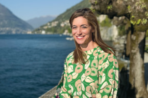 Person smiling by a lake with mountains and trees in the background.
