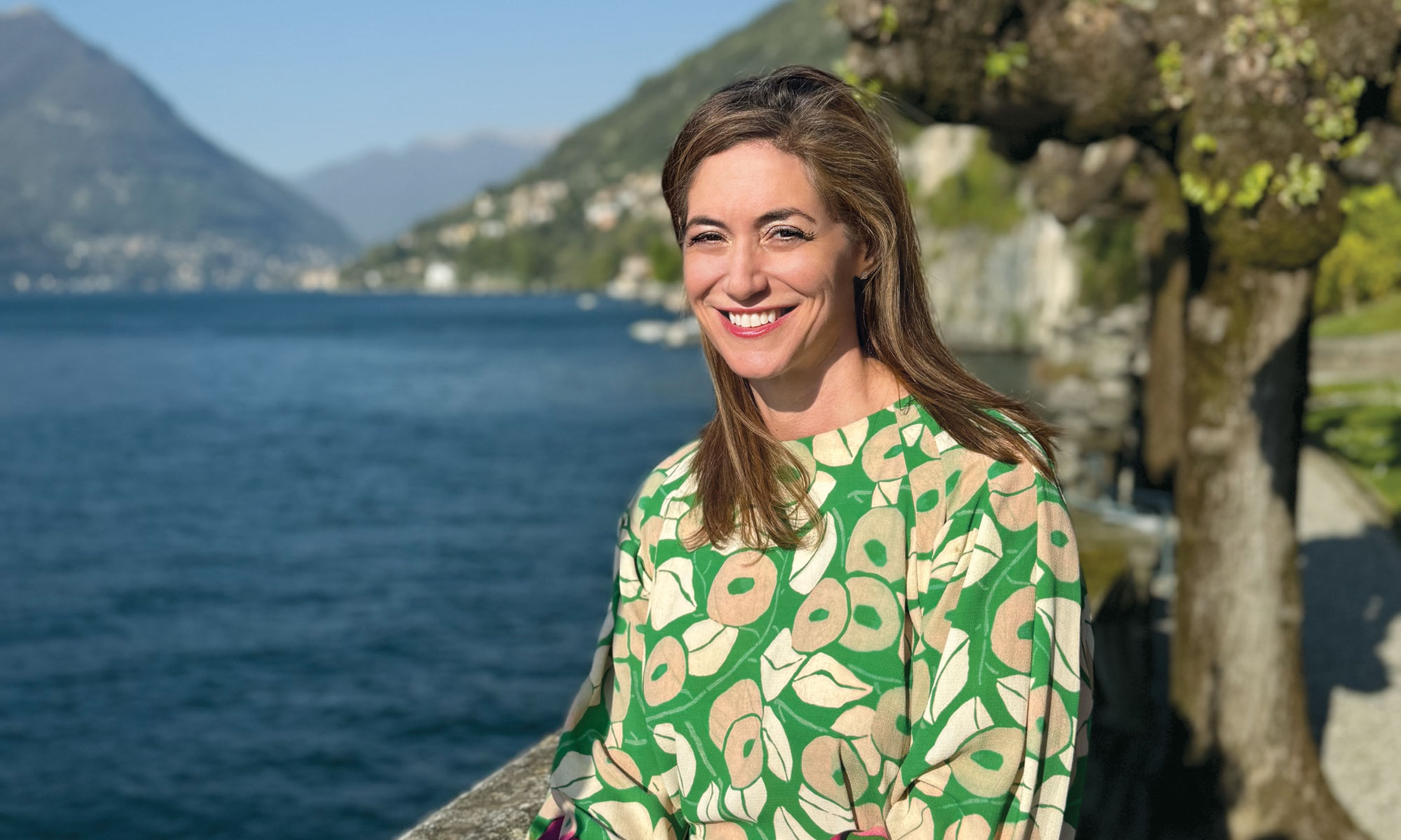 Person smiling by a lake with mountains and trees in the background.