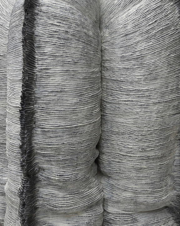 Close-up of a textured gray abstract sculpture with layered, ribbed surfaces resembling overlapping fabric or geological formations.