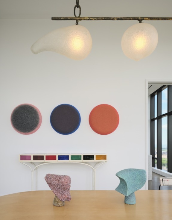 Modern art gallery with abstract sculptures on table, colorful circular wall pieces, and two pendant lights above.