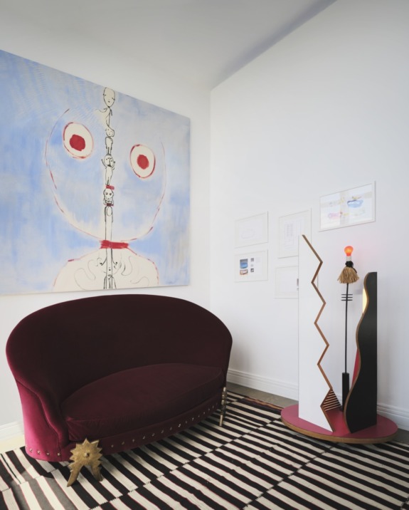 Abstract art above a maroon couch, modern geometric lamp, and striped rug in a contemporary living room setting.